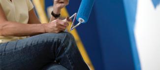 Una mano sujentando un rodillo con pintura azul enfrente de una pared pintada de azul.